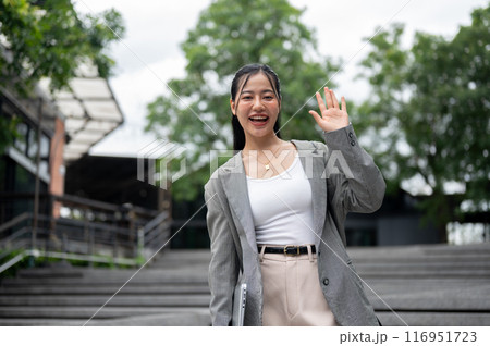 Cheerful Asian businesswoman is waving her hand and smiling at the camera while standing in the city Cheerful Asian businesswoman is waving her hand and smiling at the camera while standing in the city 116951723