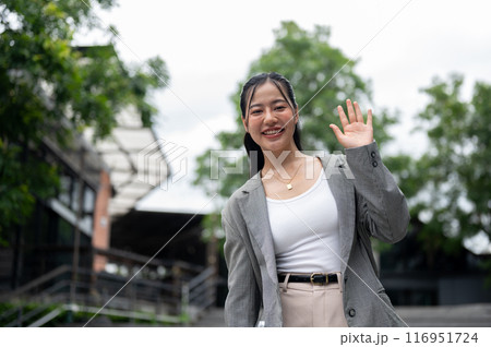 Cheerful Asian businesswoman is waving her hand and smiling at the camera while standing in the city 116951724