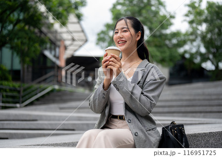 A happy Asian businesswoman is having her morning coffee while sitting on the stairs in the city. 116951725