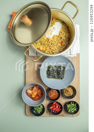 A pot of instant noodles with a lid on top of a wooden board with several bowls of food 116953294