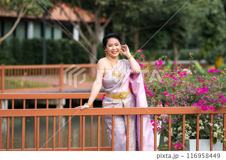 A woman in a traditional Thai costumes is standing on the wooden bridge while being taken a portrait photo shooting. A woman in a traditional Thai costumes is standing on the wooden bridge while being taken a portrait photo shooting. 116958449