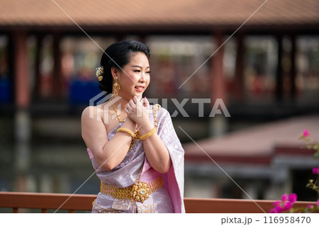 A woman in a traditional Thai costumes is standing on the wooden bridge while being taken a portrait photo shooting. 116958470