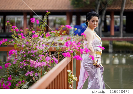 A woman in a traditional Thai costumes is standing on the wooden bridge while being taken a portrait photo shooting. A woman in a traditional Thai costumes is standing on the wooden bridge while being taken a portrait photo shooting. 116958473