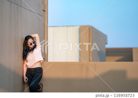 A cute Asian woman stands up and raise elbow up against the wall and wears blue sunglasses at the rooftop of the building at sunset time. 116958723