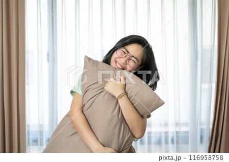 Glasses beautiful Asian woman is hugging her pillow and smiling to the camera in the bedroom with a pastel green - brown color theme. 116958758