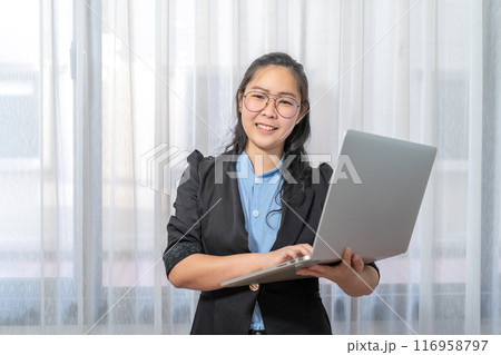 Laptop in Asian woman's hand, stands in front of window at private bedroom 116958797