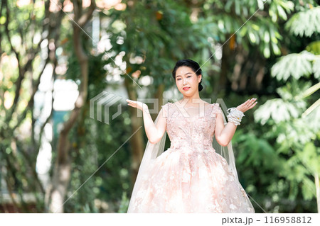 Asian girl in an old rose pink color knee length fluffy dress with high heel slingback shoes and ceremonial thread on her wrist. She is brighter mood posing in the shade of tree garden. Asian girl in an old rose pink color knee length fluffy dress with high heel slingback shoes and ceremonial thread on her wrist. She is brighter mood posing in the shade of tree garden. 116958812