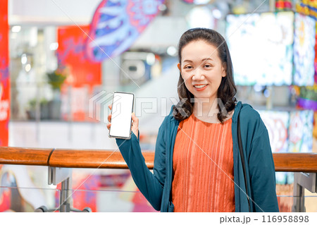 Smiling Asian Female Looks at the camera with holds smartphone in hand, Stands in Department Store Atmosphere. 116958898