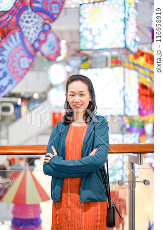 Woman wearing orange shirt and blue jacket put her hand in the pocket and smile with camera in shopping mall. Woman wearing orange shirt and blue jacket put her hand in the pocket and smile with camera in shopping mall. 116958919