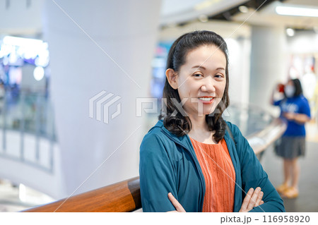 Woman wearing orange shirt and blue jacket put her hand in the pocket and smile with camera in shopping mall. 116958920