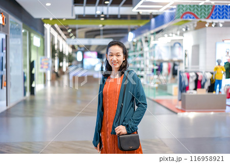 Woman wearing orange shirt and blue jacket put her hand in the pocket and smile with camera in shopping mall. Woman wearing orange shirt and blue jacket put her hand in the pocket and smile with camera in shopping mall. 116958921