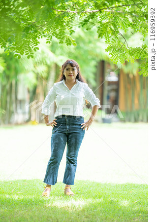 Pretty woman wearing a trusty white jeans shirt and pretty smile to the camera under the tree outside field. 116959392