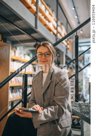 A woman in a plaid jacket sits on the stairs in a bookstore 116959404