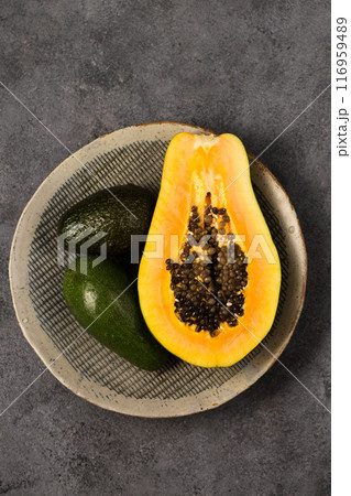 Two avocados and half a papaya on a clay plate made by hand on a gray background 116959489