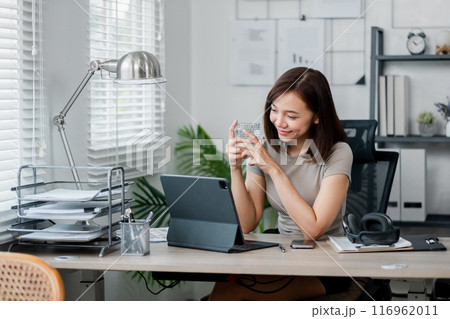 A young woman working from her home office, using a tablet and enjoying a cup of coffee. The modern workspace is bright and organized. A young woman working from her home office, using a tablet and enjoying a cup of coffee. The modern workspace is bright and organized. 116962011