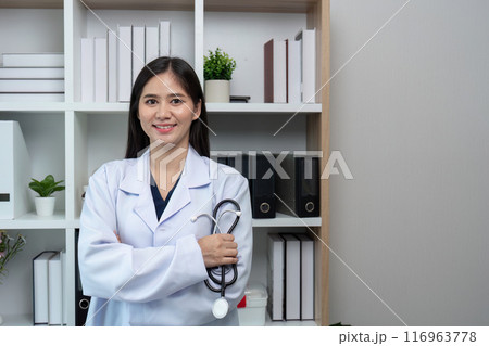 Confident Female Doctor in White Coat Holding Stethoscope in Modern Medical Office with Bookshelves 116963778