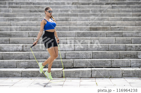 Outdoor shot of woman in sportswear exercising with skipping rope outdoors in a park. Fitness female working out with jump rope in a park 116964238