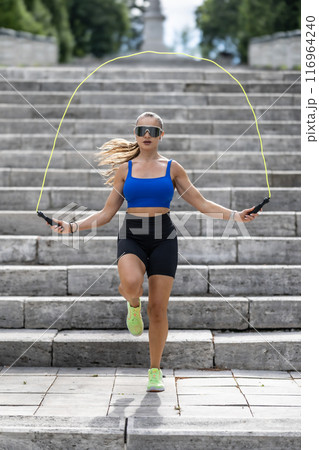 Outdoor shot of woman in sportswear exercising with skipping rope outdoors in a park. Fitness female working out with jump rope in a park 116964240