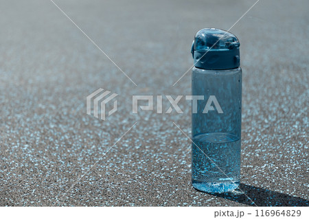 Drinking water concept. Blue refreshing bottle of water in sunny day with hard shadow on sports ground. Water balance for heat season for workout, activity and outdoor training. Close up. Copy space. 116964829