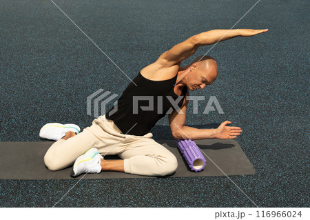 Athlete man exercising and stretching on foam roller on sports ground. Outdoor activity. Morning routine. Athlete man exercising and stretching on foam roller on sports ground. Outdoor activity. Morning routine. 116966024