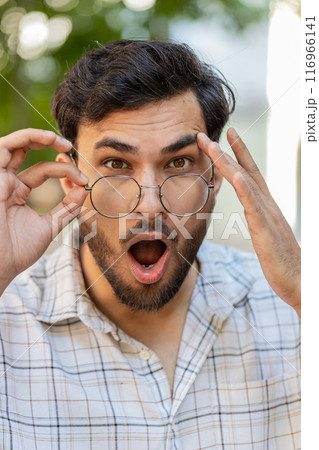 Young Indian man looking surprised at camera with big open eyes adjusting glasses on city street Young Indian man looking surprised at camera with big open eyes adjusting glasses on city street 116966141