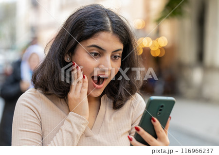 Excited happy winner Indian woman use smartphone celebrating win good message news on city street Excited happy winner Indian woman use smartphone celebrating win good message news on city street 116966237