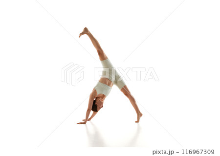 Sporty woman in athletic wear performs one-legged downward dog yoga pose demonstrating her flexibility against white studio background. Sporty woman in athletic wear performs one-legged downward dog yoga pose demonstrating her flexibility against white studio background. 116967309