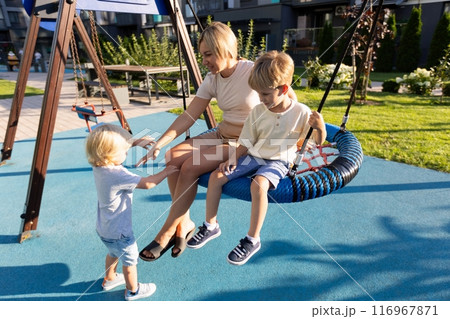 Mother Enjoys Quality Time With Two Sons Playing on a Colorful Playground Swingset in the Afternoon 116967871
