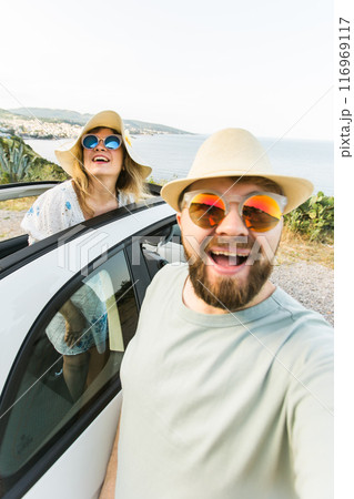 Romantic couple making selfie on smartphone camera in rental cabrio car on ocean or sea beach enjoying summer vacation together and taking picture on cellular resting near sea on weekends 116969117