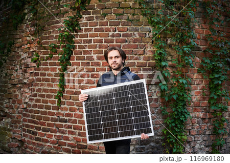 Portrait of man holding solar panel in front of old brick wall covered in green ivy. Scene blends historic charm of brickwork with modern sustainable technology. Portrait of man holding solar panel in front of old brick wall covered in green ivy. Scene blends historic charm of brickwork with modern sustainable technology. 116969180
