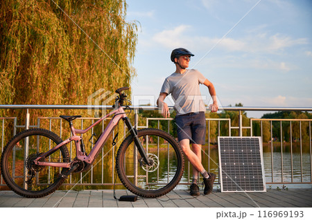 Man cyclist using solar panel for charging electric mountain bike outdoors. Guy in helmet and sunglasses stands on lakeside pathway, enjoying serene environment under clear evening sky. 116969193