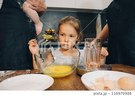 A Young Child Actively Assisting in the Family Kitchen Using Eggs and Various Cooking Tools. Happy family enjoys moments of cooking at home. A Young Child Actively Assisting in the Family Kitchen Using Eggs and Various Cooking Tools. Happy family enjoys moments of cooking at home. 116970986
