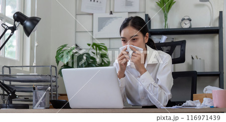 Young Professional Woman Sneezing at Work Desk, Office Environment, Sick Day, Flu Season, Health and Wellness Concept Young Professional Woman Sneezing at Work Desk, Office Environment, Sick Day, Flu Season, Health and Wellness Concept 116971439