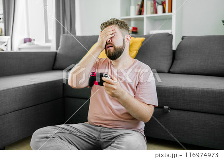 Young man spending time at home, sitting on a couch in apartment and playing arcade car video games on console. Male using controller to play street racing drift simulator. Young man spending time at home, sitting on a couch in apartment and playing arcade car video games on console. Male using controller to play street racing drift simulator. 116974973