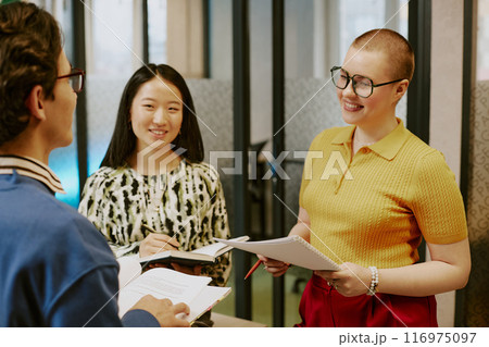 Over shoulder medium shot of cheerful young biracial women and man holding documents and notebooks standing in office and talking 116975097