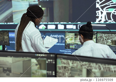 Manager offers logistics papers to indian employee in observation room, following delivery trucks on satellite navigation for routes around the city. Worker operating on CCTV public system. Manager offers logistics papers to indian employee in observation room, following delivery trucks on satellite navigation for routes around the city. Worker operating on CCTV public system. 116975509