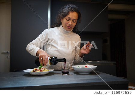 Woman pouring red wine into a glass while preparing a meal in a modern kitchen 116975659