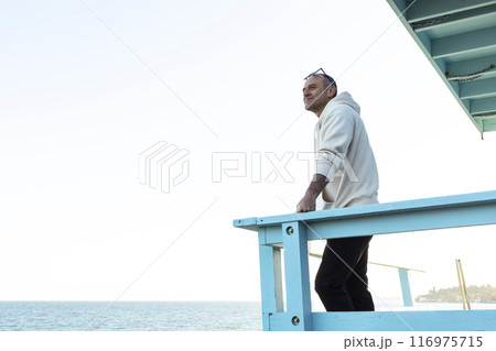 Middle Age Caucasian Man Standing Alone On Wooden Pier And Staring At Sea, Ocean At Sunny Day. Relaxed 40 Yo Male Wears Hoodie, Denim, Sunglasses. Vacation, Trip, Relaxation. Peaceful Atmosphere. 116975715