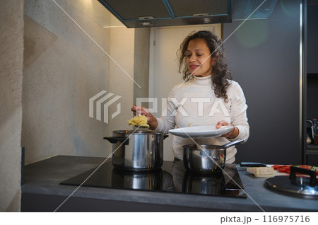 Woman cooking in modern kitchen, stirring pot on stove, and preparing meal with fresh ingredients 116975716