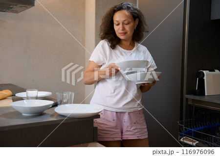 Woman loading dishwasher in modern kitchen while preparing meal Woman loading dishwasher in modern kitchen while preparing meal 116976096
