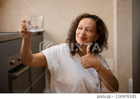 Woman in white t-shirt giving thumbs up while inspecting a clear glass in the kitchen Woman in white t-shirt giving thumbs up while inspecting a clear glass in the kitchen 116976185