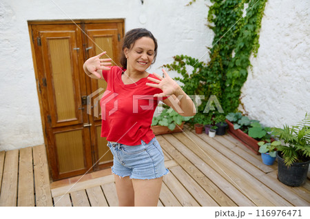 Happy woman dancing outdoors on a wooden deck with rustic door and green plants background 116976471