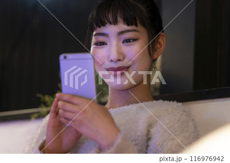 スマートフォンを操作する笑顔の若い女性　夜の自宅　イメージ 116976942