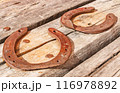 Two old vintage and rusty horseshoes are nailed to a dried and cracked piece of wood. 116978892