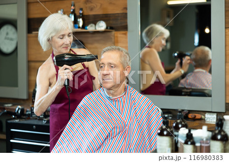 Female barber using hairdryer to style hair to elderly client in barbershop Female barber using hairdryer to style hair to elderly client in barbershop 116980383