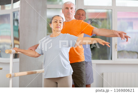 Pensioners of different ages exercise near ballet barre at group training session in two position Pensioners of different ages exercise near ballet barre at group training session in two position 116980621