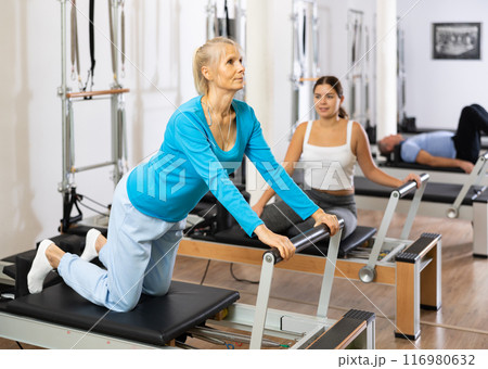Two sporty focused young mature women in sportswear performing Pilates exercise for back on reformer bed machine in gym 116980632