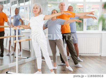 Group of positive aged women and men standing in row together while doing ballet dance moves, dancers standing in 2nd position of ballet stance in fitness room Group of positive aged women and men standing in row together while doing ballet dance moves, dancers standing in 2nd position of ballet stance in fitness room 116980918