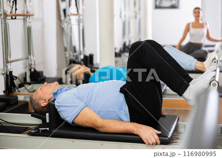 Elderly male pensioner lying down on bed of Pilates reformer bed machine with legs up, working out and recovering in gym Elderly male pensioner lying down on bed of Pilates reformer bed machine with legs up, working out and recovering in gym 116980995
