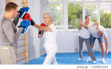 Mature woman with her trainer at gym. Woman wearing boxing gloves exercise and punch to pads for boxing Mature woman with her trainer at gym. Woman wearing boxing gloves exercise and punch to pads for boxing 116981161
